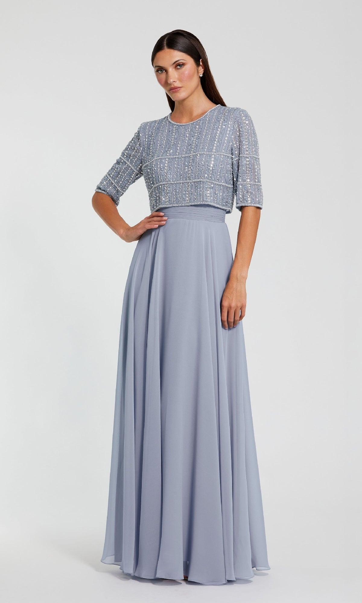 A woman wears the Mac Duggal 9180 Short Sleeve Long Formal Dress, featuring a beaded bodice and flowing light blue chiffon skirt, standing against a plain light background.